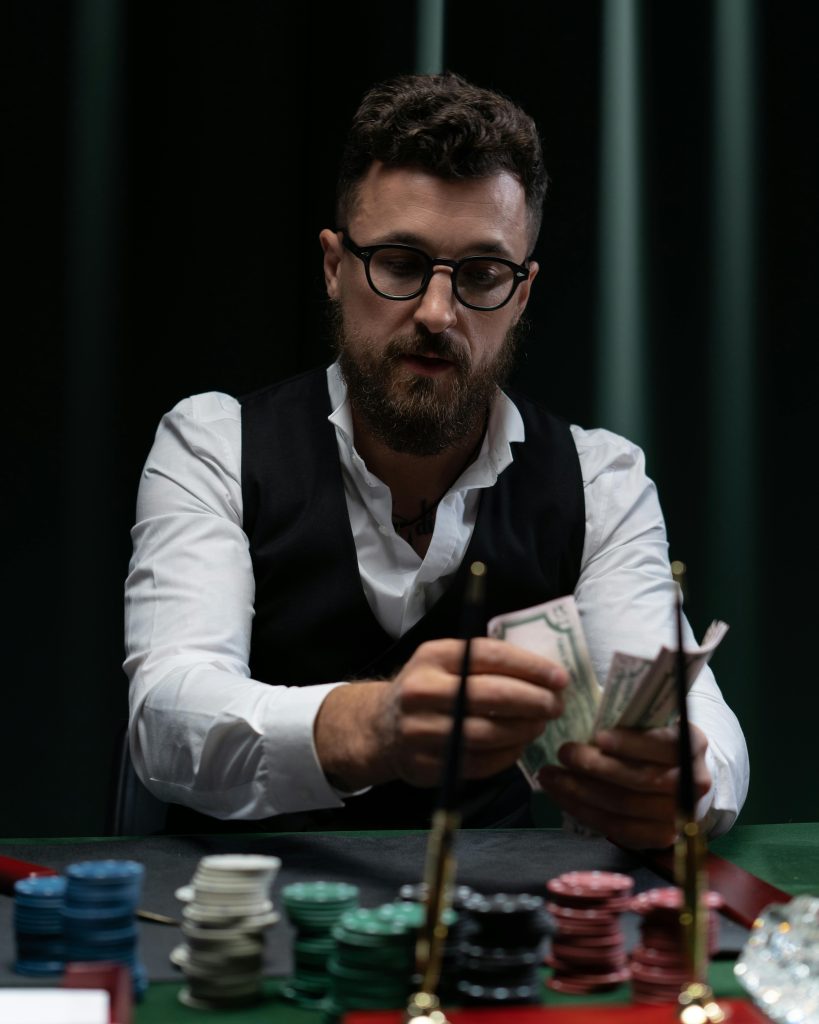 Bearded man in glasses counting money at a poker table with poker chips in view.