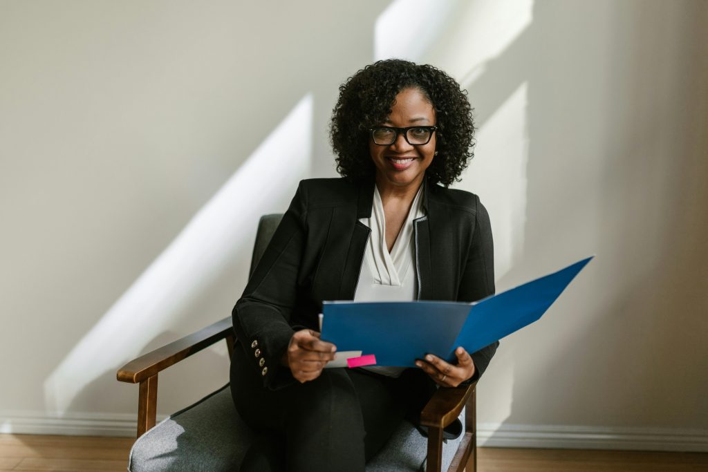 Professional African American woman with glasses and curly hair holding a blue folder indoors.